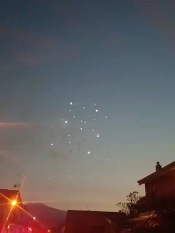 The video captures a sequence of fireworks exploding in the dusky sky. The camera is positioned looking upwards from a street level, with buildings and a street light visible in the foreground, silhouetted against the twilight. The first few seconds show smaller, less distinct bursts of light, followed by larger, more vibrant explosions of fireworks in various colors, including white and red. The fireworks create a dazzling display against the deepening blue of the sky. The overall atmosphere suggests a celebratory event taking place as dusk settles.