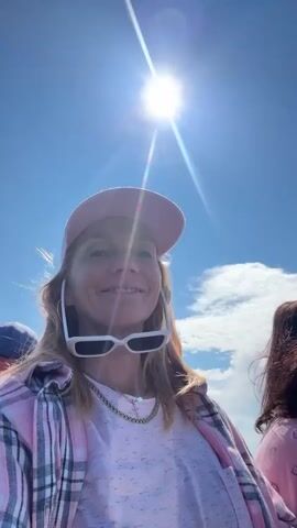 A woman in a pink baseball cap and a pink and white plaid shirt smiles at the camera, holding her sunglasses on top of her cap. She is on a boat with two other people. A man in a blue baseball cap and an orange life vest is visible to her left, and another woman wearing sunglasses and a pink shirt is to her right, looking out at the water. The sun is shining brightly in a clear blue sky with a few clouds. The water is calm and reflects the sky.
