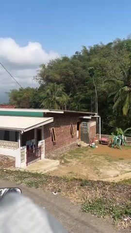 The video shows a view from a vehicle traveling on a dirt road in a rural area. The road is lined with houses, some of which appear to be under construction or renovation. The houses are made of brick and have tile roofs. There are also trees and vegetation in the background, including some palm trees and bamboo. The sky is blue with some clouds. The camera is focused on the scenery outside the vehicle, and it is unclear what is happening inside the vehicle.