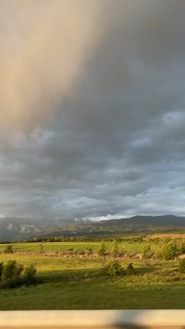 The video captures a scenic landscape from a moving vehicle, likely during the late afternoon or early evening as suggested by the varied lighting. In the foreground, green fields extend towards a distant mountain range, some peaks of which are covered with snow. The sky is dramatic, dominated by dark, heavy storm clouds on the right, while the left side shows lighter, possibly sun-kissed clouds. A yellow car briefly appears on a road cutting through the fields. A white fence is visible in the immediate foreground, indicating the perspective from a car or train. Lush green trees and vegetation are scattered across the landscape. The scene exudes a sense of natural beauty and vastness, with the contrasting light and dark clouds adding to the dramatic atmosphere. Music is audible in the background. The location is Barakovo, Bulgaria.