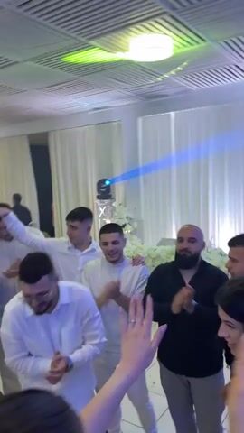 The scene takes place in a banquet hall with a group of people dancing and clapping. The room is decorated with white curtains and floral arrangements. Most of the men are dressed in white, while one man is wearing a black suit with a bow tie. There is a woman in a white dress and a man in a black shirt in the group. The camera moves around the room, capturing different angles of the celebration. A drum set is visible in the background. A man in the center of the group is wearing all white and clapping enthusiastically. A lighting rig is pointed down from the ceiling. Overall, the atmosphere is festive and lively.