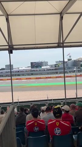 The video shows a view of a race track from under a white tent. In the foreground, the backs of several people are visible, sitting in blue seats. Most of them are wearing red shirts with a Ferrari logo and "hp" printed on them. A race car with a purple and black design is visible on the track in the distance, with a large screen showing a race, and grandstands in the background. The track has green and white markings. The weather appears to be overcast.
