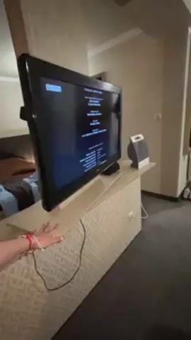 A person stands in a room with dark carpet and patterned wallpaper, near a TV stand. The TV displays the end credits of a movie. A small white heater sits next to the TV. A person's hand with a bracelet is visible near the edge of the TV stand. A doorway is visible in the background.