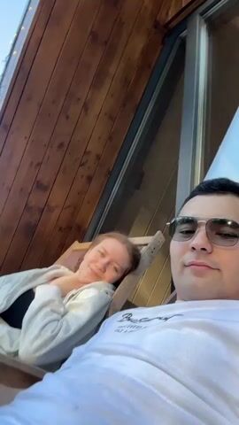 A young couple is relaxing in deck chairs outdoors. The woman on the left has reddish hair and wears a black top under a grey cardigan. She rests her head on her hand and smiles. The man on the right wears sunglasses and a white t-shirt. They are positioned against the side of a wooden structure with an angled roof and a large window. There are small lights lining the roof. They are looking directly at the camera.