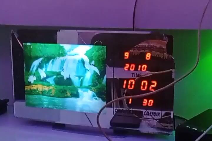 The video shows a homemade device consisting of a small screen displaying a looping image of a waterfall next to a digital clock that shows the date and time as September 8, 2010 and 10:02.  Wires connect the device to other components which are partially visible.  The device sits on a surface under ambient lighting with a purple hue.  Music plays in the background.