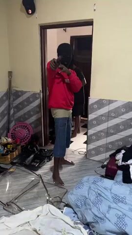 Two barefoot men are in a messy room. One, wearing a red sweatshirt, blue jean shorts, and a black ski mask, aims a handgun at the camera. The other man, wearing a black t-shirt and dark shorts, stands behind the first. A doorway is visible behind them. The room contains a broken plastic chair, a pink fan, shoes, luggage, and piles of clothes. The walls are a light yellow, with a decorative, patterned tile border. A security camera is visible in the upper left corner.