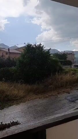 The video shows a brief outdoor scene on a cloudy day. In the foreground, there is a wooden railing or ledge. Beyond that, a wet asphalt road is visible, with dry grass and some bushes bordering it. In the background, there are houses with tiled roofs, a large green tree, and mountains in the distance. The sky is partly cloudy with patches of blue. Small white particles appear to be falling from the sky, suggesting rain or possibly snow, though the overall environment seems more temperate. There are no people or vehicles clearly visible.