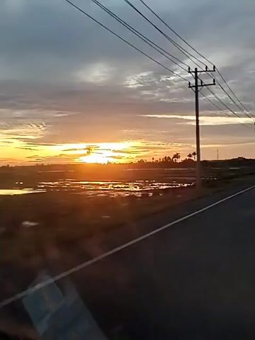 The video captures a sunset scene from a moving vehicle, looking out the side window. A paved road with a white shoulder line runs diagonally across the lower right of the frame. To the left of the road, a wide, flat landscape appears, possibly a wet or flooded field, reflecting the bright golden light of the setting sun which is low on the horizon. The sky above is cloudy, with a mix of dark gray and lighter clouds. Several utility poles with power lines stretch across the upper and middle sections of the frame, receding into the distance. Sparse trees, some resembling palm trees, are visible in the distance on the horizon. The overall impression is a serene, rural evening.