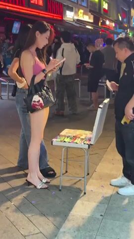 A woman wearing a pink top and short shorts is standing in the street with a woman next to her. The woman in shorts is looking at her phone, while a man on her right is looking at a table with colorful items. The table is silver and folds up like a chair. In the background, people are walking on the sidewalk in front of various shops and restaurants. The buildings have bright neon signs. The sidewalk is paved with hexagonal tiles.