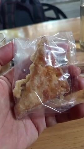 A hand holds a triangle-shaped fried food item packaged in clear plastic. The food has a golden-brown color and uneven edges, appearing crispy. The hand is fair-skinned with visible veins, suggesting an adult. The food is presented against a wooden table, with glimpses of a backpack and a glass of liquid in the background. The scene is well-lit.