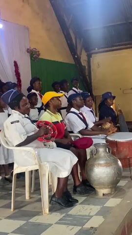 The video shows a group of people performing music inside a building. Most are wearing uniforms with a navy blue hat. They stand behind a row of four people sitting in chairs. The people sitting down play drums, shakers and a tambourine. The group is on a stage, which is painted with checkered squares. A large, silver urn-like object is between the chairs and the drum. A sign is behind the group, and it appears to be religious. The light inside is bright.