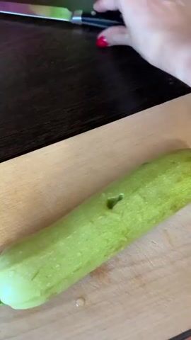 This close-up video shows a person's hands preparing a zucchini. The zucchini is light green and lies on a wooden cutting board. A knife is used to cut the stem end off the zucchini. The background is blurred but appears to be a dark wooden surface. The activity suggests food preparation, likely for cooking. The lighting is bright, indicating it is daytime. No text is visible in the media.