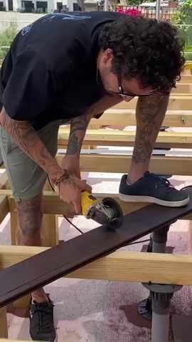 A person with tattoos on their arms and legs is using an angle grinder to cut a dark-colored board. They are wearing a black t-shirt and green shorts, and black sneakers. The board is resting on a wooden frame that appears to be part of a deck or platform, supported by adjustable pedestals. The background shows a building and some greenery. The sun is shining, suggesting an outdoor setting.