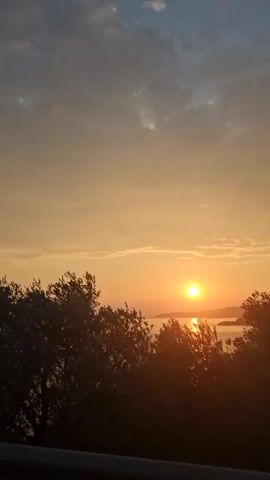This video shows a sunrise over a body of water. The sun is a bright orange orb partially obscured by trees in the foreground. The sky is a gradient of orange and blue, with some clouds. The water is calm and reflects the colors of the sky.
