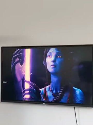 A flat screen television is mounted on a white wall. The television is displaying a scene from the movie Avatar. The scene transitions from a close-up of a blue-skinned alien with braided hair looking at a glowing purple object to a wider shot of two figures, one human and one alien, sitting in a fantastical, glowing forest. The overall color palette of the scene is dark and mystical, with vibrant bioluminescent flora. The television has a black frame and a visible logo at the bottom center. A black cable extends from the bottom right of the television.