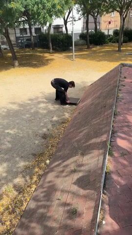 The video shows a person in a black t-shirt and black pants wearing black shoes with white soles. They are in an outdoor area with a dirt and gravel ground covered in yellow leaves. To the right of the person is a slanted red brick structure. The person is interacting with a flat, dark rectangular object placed on the ground, partially against the brick structure. The person bends down and places their right foot onto the object, then places their left foot on it, standing up fully. They then place their hands on their hips and look off to the side. The camera is positioned at an angle looking down at the scene.