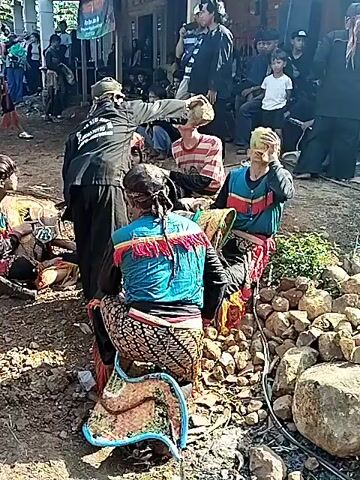 The video shows a group of people gathered in what appears to be a rural or village setting. Most of the people are wearing traditional Indonesian clothing, including colorful jackets with fringe, batik pants, and headbands. Some are holding coconut halves to their faces. In the background, there is a building with a porch and a group of spectators watching. A person in a mask is performing in the background. The ground is uneven and rocky. The scene suggests a cultural event or traditional performance taking place.