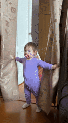 Toddler in purple onesie stands by curtains in Krapivka