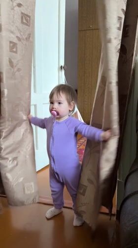 Toddler in purple onesie stands by curtains in Krapivka