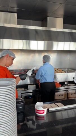 The video captures a dynamic scene within a commercial kitchen, likely a restaurant or butcher shop, located in Silivri, Turkey. Three individuals, all adhering to hygiene standards by wearing hairnets and gloves, are actively engaged in preparing food.

In the foreground to the left, a young man dressed in an orange long-sleeved shirt is holding what appears to be tongs and a small paper item, possibly a receipt or an order. He seems to be either preparing an item for delivery or checking an order.

Behind him, two other kitchen staff members are working at a large, professional-grade stainless steel grill. The person in the center wears a light blue shirt and black pants, while the individual to the right wears a white shirt and black apron. Both are facing away from the camera, busily tending to numerous food items laid out on the grill, which resemble meat patties or sausages. A clear label with "SUCUK" (Turkish sausage) is visible on the equipment below the grill, hinting at the type of food being prepared.

The kitchen is characterized by its clean, industrial design, featuring extensive stainless steel surfaces, including the grill area, backsplashes, and overhead ventilation. On the sleek, black countertop in the immediate foreground, there are stacks of white plates, a black Xprinter device, and a white disposable container with red text. The text on the container, visible as a reflection, indicates "KASAPLI KASAP" (Butcher's Butcher), further suggesting the establishment specializes in meat products. The ambient sound includes Turkish speech, with words like "köfte" (meatballs/patties) and "karışık" (mixed) being mentioned, reinforcing the culinary context.