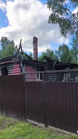 The video shows the exterior of a damaged building with a partially collapsed roof, blackened wood, and a brick chimney. A dark brown fence runs in front of the building. In the background, there are trees and a bright blue sky with white clouds. The camera pans to show a gravel path leading towards a gate. Further down the path, a person wearing a white hoodie and grey sweatpants is walking away from the camera. A small dog on a leash is walking beside the person. The video also shows a white house with a metal roof and a ladder leaning against the side.