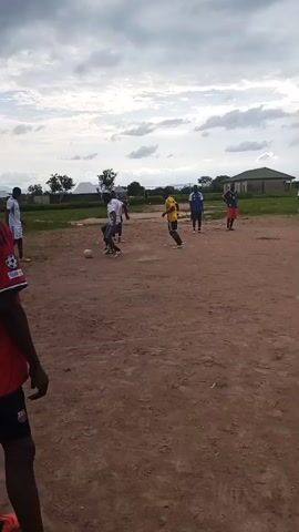 The video shows a group of young people playing soccer on a dirt field. The sky is cloudy. In the foreground, a person in a red shirt is facing away from the camera. In the background, several other individuals are visible, some in athletic wear. One person is kicking a soccer ball, while others are running or standing nearby. Buildings and trees are visible in the distance. The overall scene depicts an informal soccer game.