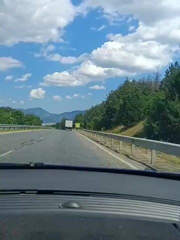 The video shows a first-person perspective from inside a car traveling on a highway. The road is paved with multiple lanes, separated by white lines. On the right side of the road, there is a metal guardrail and a steep incline covered in dense green trees. In the distance, mountains are visible under a bright blue sky with scattered white clouds. A white truck is visible in the distance on the highway. A yellow road sign with multiple warning symbols and text is posted on the right side of the road, just past the guardrail. The interior of the car, including the dashboard, is visible in the foreground. The video is in color and appears to be captured during daylight hours.