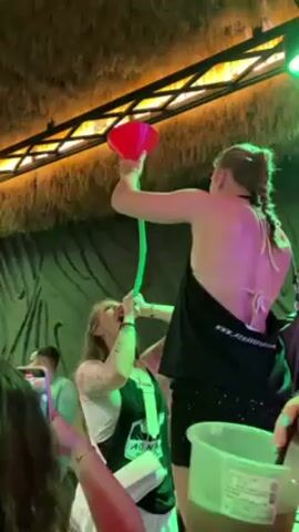 The video shows a young woman with blonde hair and braids holding a red funnel attached to a green tube. The tube is in the mouth of another young woman with tattoos who is drinking from it. The first woman is standing and wearing a black top that is partially covering a white bathing suit top. She is also wearing a sparkly black skirt. There is a large container of liquid next to her. The woman who is drinking is wearing a white shirt and black tank top. There are other people surrounding the women, some of whom are holding phones and recording. The video appears to be taken inside of a bar with green lights and jungle-themed decorations.