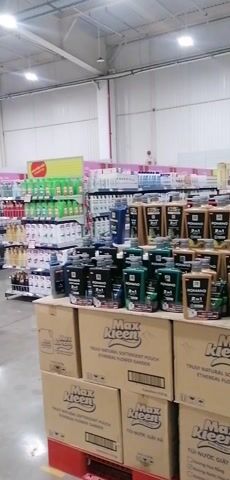The video shows a walk-through of a large supermarket or hypermarket. The setting features wide aisles with concrete floors and a high ceiling equipped with fluorescent lighting. Various products are neatly displayed on shelves and stacked on pallets throughout the store.

In the initial frames, an aisle with cleaning supplies and personal care items is visible, including large bottles and boxes labeled "Max Kleen." As the camera moves, it pans across different sections, showcasing packaged goods like canned drinks (including "Red Bull" and "MILO"), bottled water ("Aquafina"), and other boxed products. There are also sections with household items like dishes and bowls. The store appears spacious and well-organized.

Notable text includes "Max Kleen" and "Aquafina" on product packaging. Overhead signs display Vietnamese text such as "Thu Ngân" (Cashier), "Thêm cách Hiện Đạt" (More modern ways), and "Hàng Tươi Sống" (Fresh food). Red circular signs with numbers "11, 10, 9, 8, 6" are also visible, likely indicating aisle or checkout numbers. The overall impression is that of a bustling retail environment in Ha Tu Ward, Vietnam.
