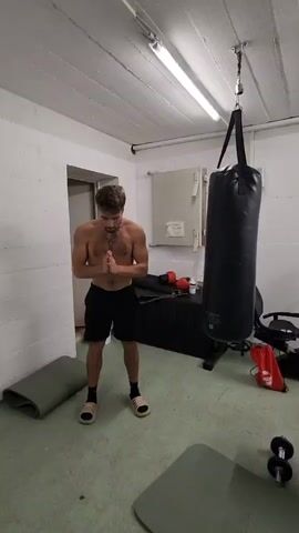 A shirtless man in black shorts and tan sandals practices boxing moves in what appears to be a home gym. He starts by bending over with his hands on his knees, then straightens up into a boxing stance, and begins throwing punches at a black punching bag hanging from the ceiling. The gym is in a basement-like room with white walls and a low ceiling with exposed pipes. Other items in the room include exercise mats, dumbbells, boxing gloves, and a cabinet.