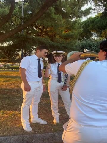 A man in a white shirt, dark tie, and white pants stands on the left, looking at the camera and smiling. He is wearing sunglasses. Next to him, a person in a similar uniform holds drumsticks. In the foreground, a person in a white shirt and pants plays a brass instrument. The scene is outdoors, with trees and grass visible. The lighting suggests it is late afternoon or early evening.