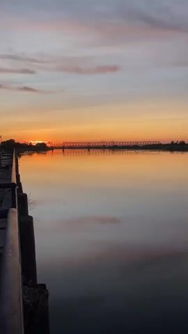 A vertical shot captures a serene sunset over a body of water. The sun, a vibrant orange orb, is setting on the horizon, casting a warm glow that reflects beautifully on the calm water's surface. A long, thin bridge stretches across the water in the distance, silhouetted against the colorful sky. The sky itself is a gradient of soft blues and oranges, with wispy clouds adding texture. In the foreground, the edge of a pier or boardwalk is visible on the left, with a dark railing. The water is dark and still, mirroring the colors of the sky and the distant bridge. The overall scene evokes a sense of peace and tranquility.