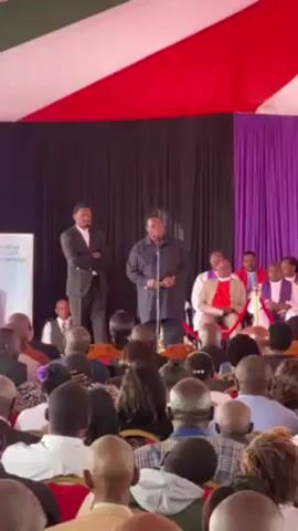 The video shows a man speaking into a microphone in front of a seated audience. The speaker is wearing a dark jacket, and there is a man standing behind him to his left in a dark suit. Behind the speaker, there is a group of individuals who appear to be clergy, wearing religious robes and seated in chairs. To the left of the stage, there is a sign with text and an image of a person, although the details are not fully discernible. The audience consists of people of diverse ages and appearances. They are all facing the stage. The setting appears to be a formal event, possibly a memorial service or a public gathering, held under a tent decorated with red, white, and green fabric.