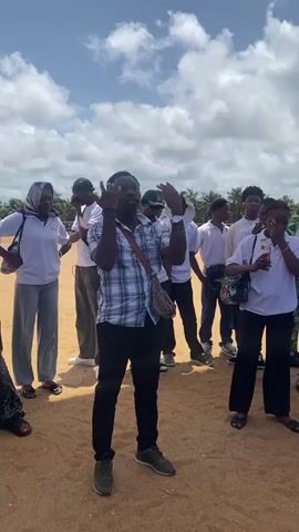 The video shows a man in a plaid shirt addressing a group of young people and adults on a sandy ground with trees visible in the background. The people in the group are listening attentively. The sky is partly cloudy. Several people are wearing white shirts and carrying bags. The man in the plaid shirt is gesturing with his hands as he speaks.