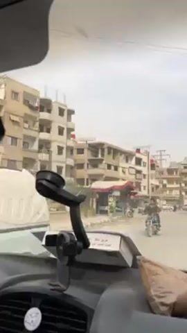 The view is from the inside of a vehicle, likely a car, looking out through the windshield. The car is at an intersection in what appears to be a Middle Eastern town. Several buildings of varying heights are visible, appearing to be made of concrete or similar materials, some looking incomplete or damaged. The sky is overcast, with a general sense of dustiness in the air. 

In the foreground, inside the car, there is a dashboard with a phone mount and some loose items. Outside, people are seen riding a motorcycle. There is also car and other motorcycles going through the intersection. There are also electric poles and wires visible in the background.
