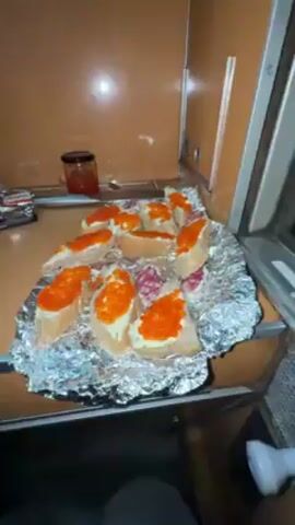 The video shows a man pouring champagne into a cup inside a train cabin. The cabin has a shelf with food items, including sandwiches with caviar and salami. The man is wearing a beige hoodie and sitting on a seat. There is a yellow puffer jacket hanging on a hook. The walls of the cabin are wood-paneled.
