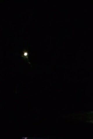 The video captures a first-person perspective driving at night on a dark asphalt road. The headlights of the vehicle illuminate the road ahead, revealing white lane markings. In the distance, other vehicles can be seen with their headlights on. The sky is black, indicating nighttime. There are some faint specks visible, possibly rain or dust on the windshield. The overall environment appears to be a rural or unlit highway.