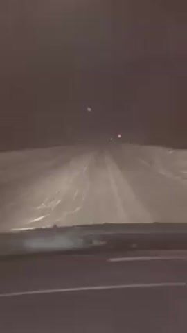 The video shows a driver's perspective of driving on a dark, snowy road at night. The road is covered in snow, and snow is falling heavily, obscuring visibility. The headlights illuminate the road ahead, making the falling snow visible. A triangular warning sign is visible on the right side of the road, also covered with snow. The dashboard of the car is visible at the bottom of the frame. The overall impression is that of a hazardous driving condition due to the heavy snowfall and poor visibility.