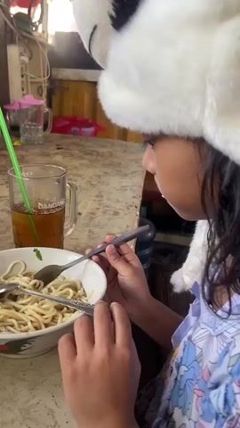 A young person wearing a panda hat is eating noodles. They are sitting at a table in what appears to be a casual eatery or home. A glass of iced tea with a green straw is next to the bowl of noodles. A smartphone is propped up, playing a video of several people in orange outfits. The table surface is speckled brown and beige. Other items on the table include a small container with a lid, a blue utensil holder with red and silver utensils, and some green vegetables. The background shows a cluttered area with electrical outlets and various objects.