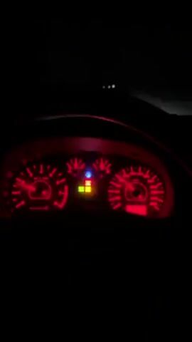 The video displays the dashboard of a car in the dark, highlighting the red illumination of the speedometer, tachometer, and other gauges. A blue light and two yellow lights are also illuminated. The camera remains static, focusing solely on the dashboard. There are some exterior lights visible through the front windshield. The primary action consists of the car's dashboard lights being visible in the dark. There are no visible people or interactions beyond the implied operation of the vehicle.