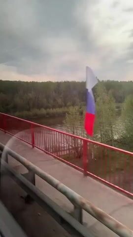 A view from a moving vehicle crossing a bridge in Russia. A Russian flag attached to the bridge’s red railing whips in the wind. A wide river flows beneath the bridge. Lush green trees line the riverbanks. The sky is overcast with grey clouds.