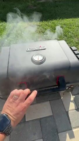 The video shows a person opening a black Char-Broil grill. Smoke billows out as the lid is lifted, revealing two large steaks cooking on the grates. The steaks appear to be thick-cut, possibly tomahawk steaks, with a bone protruding from one of them. The grill is outdoors, with green grass and a paved patio visible in the background. The sunlight suggests it is daytime. The person's hand, wearing a watch on the left wrist, is visible as they lift the grill lid. A thermometer on the grill lid indicates the temperature.