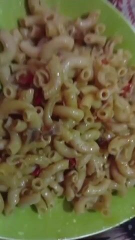 The video shows a close-up of a green, heart-shaped plate filled with cooked macaroni. The macaroni appears to be coated in a light-colored sauce with flecks of red pepper and what might be onion. Two silver forks rest on the plate. The background is slightly out of focus, revealing what appears to be a patterned surface. The camera zooms in slightly on the macaroni.