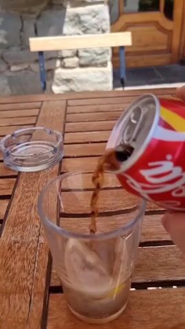 A hand is pouring Coca-Cola from a red can into a clear glass. The glass contains a small amount of dark liquid and some ice at the bottom. There is a glass ashtray to the left of the glass, on a wooden table. In the background, there is a stone wall and a wooden bench. The scene appears to be outdoors.