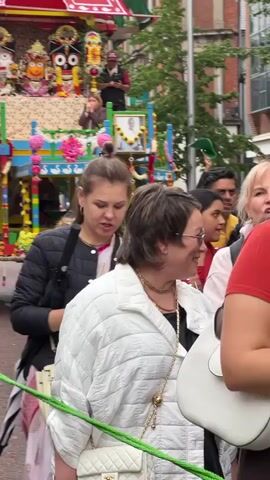 A religious procession is underway on a street. In the background, a brightly decorated chariot is visible, adorned with figures and flowers, and carrying several people, including a woman and a man in a red shirt. A portrait of a man in a white outfit is also displayed on the chariot. In the foreground, a woman in a black puffer jacket is looking at her phone, holding a white handbag and a black umbrella. Beside her, another person in a white quilted jacket is seen from the side, smiling and looking forward. A group of people, including men, women, and children, are walking behind them. One man in a bright yellow safety vest is holding a green rope. The environment appears to be an urban street with buildings visible in the background. The overall atmosphere seems festive and communal.