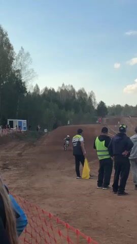 The video shows a motocross race. The track is dirt and has several jumps. A rider on a dirt bike is visible in the distance, jumping over a mound. Several people are standing on the side of the track, watching the race. One person is holding a yellow flag. There are trees and a blue and white structure in the background. The sky is clear and blue.