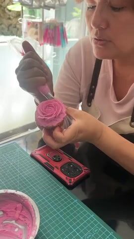 A woman with reddish-brown hair and bangs is shown from the chest up, looking down and concentrating on decorating a cupcake. She is wearing a light pink shirt and a dark brown apron. In her right hand, she holds a pink piping bag with a star-shaped tip, from which she is piping pink frosting onto the cupcake to create a rose design. Her left hand supports the cupcake. In the foreground, a blue cutting mat is visible, with a white bowl containing pink frosting to the left. A red phone case with a metallic design is placed on the cutting mat to the right of the cupcake. The background appears to be a display shelf in a store, with various items, some appearing to be cake decorating supplies like piping bags and cutters, visible. The lighting is bright, suggesting an indoor setting.