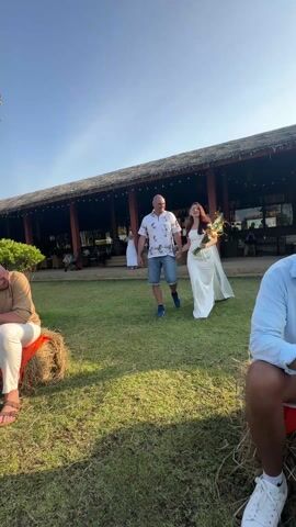 The video shows a wedding procession outdoors on a sunny day. A woman in a white satin dress with a long veil and a bouquet of flowers is walking hand-in-hand with a man in a patterned shirt and shorts. They are walking across a grassy lawn towards a building with a thatched roof. Several people are seated on hay bales on either side of the path, watching them.  The scene appears to be a relaxed and informal wedding celebration. String lights are visible in the background.