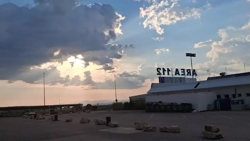 The video shows a parking lot with a building labeled "AREAIS" in the background. The sky is filled with clouds, and the sun is shining through them. There are several light poles in the parking lot. Some rocks are scattered around on the asphalt.  There are also several green bins against the building. There are cars parked in the background, further away from the building.