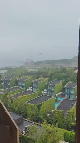 The video shows a view from what appears to be a metal tower overlooking a resort or entertainment complex on a foggy day. The structures in the foreground are buildings covered in ivy with small, private swimming pools. As the camera pans upward along the metal structure of the tower, the view reveals a dome-shaped building and what looks like a rollercoaster in the distance. The visibility is low due to the fog.